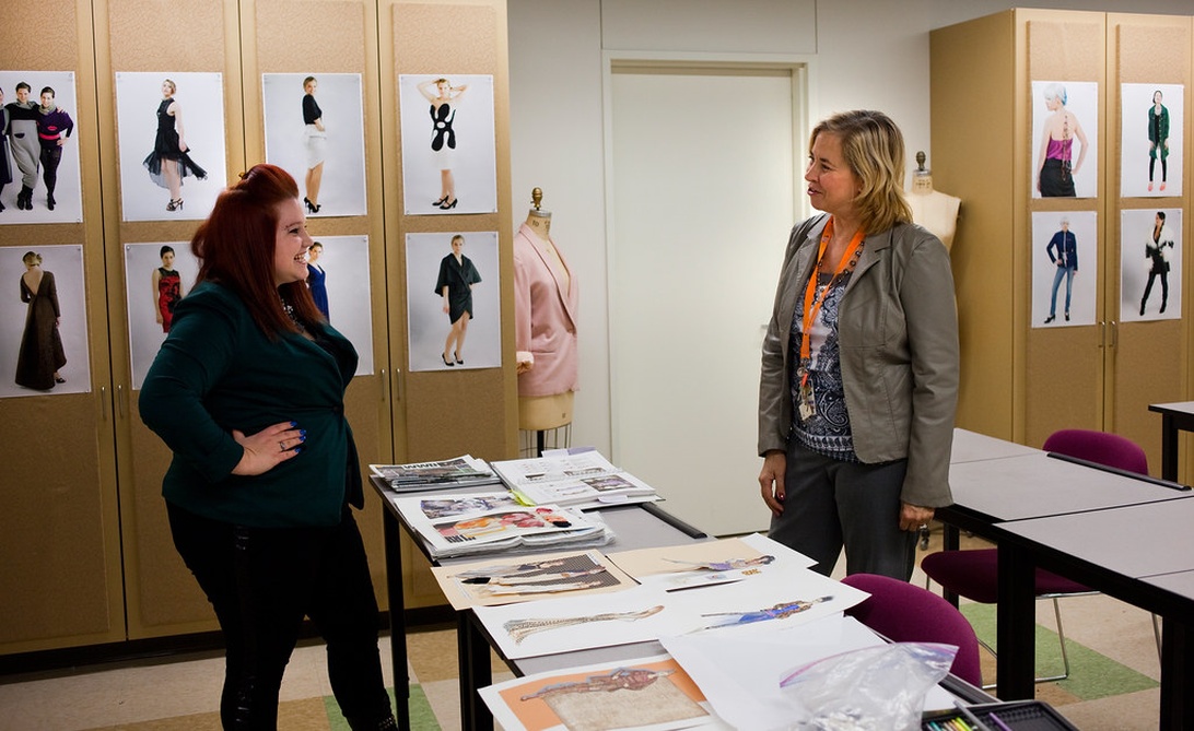 Een modeontwerpstudio waar twee vrouwen ontwerpen bespreken, met schetsen en een paspop op de achtergrond.